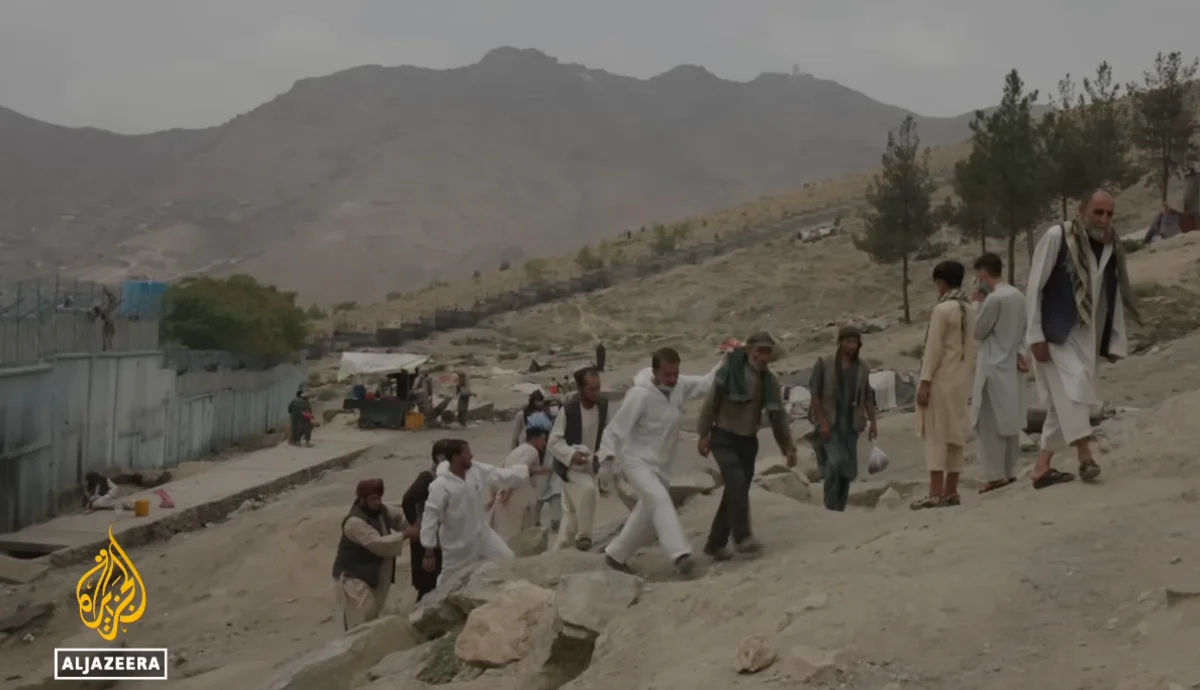 A group of homeless men walk up a steep, rocky hill near a fence in a barren landscape, while being escorted by Ibn Sina staff dressed in white PPE clothing. Mountains are visible in the background under an overcast sky.