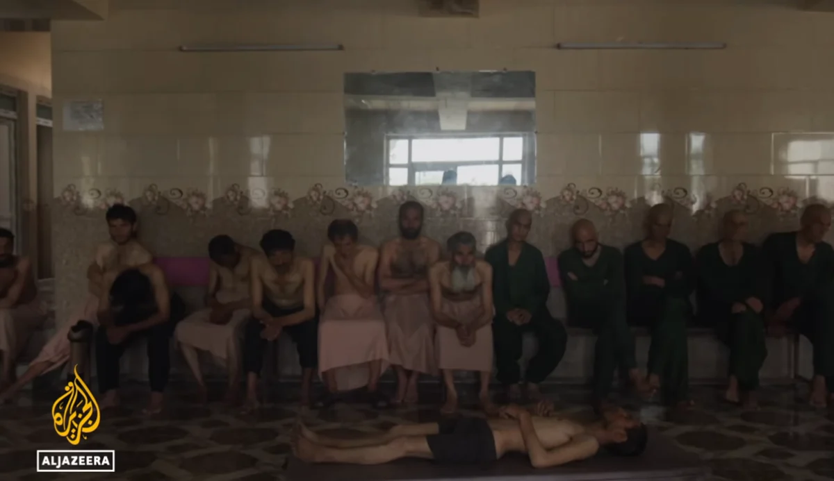 Several shirtless and robed men sit on benches inside a tiled room, watching another man lie flat on a table in the foreground.