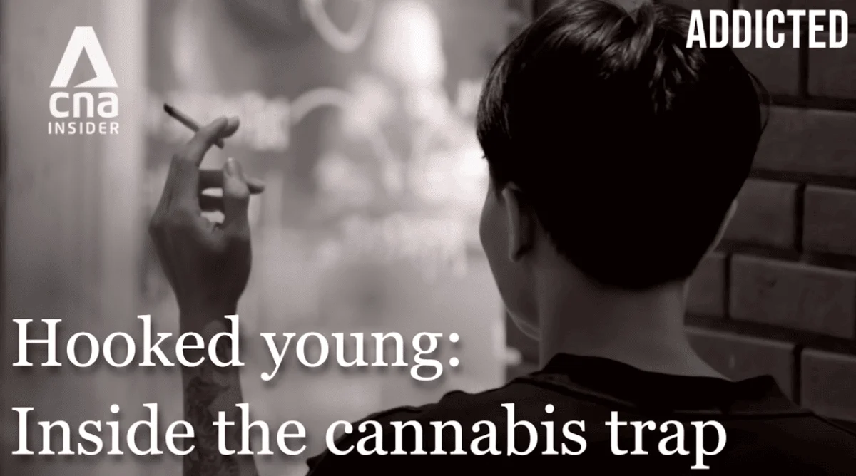 A young man with his back facing the camera and his arm slightly raised to show his hand holding a cannabis joint between his fingers, with text overlay reading "Hooked Young: Inside the Cannabis Trap" and "ADDICTED".