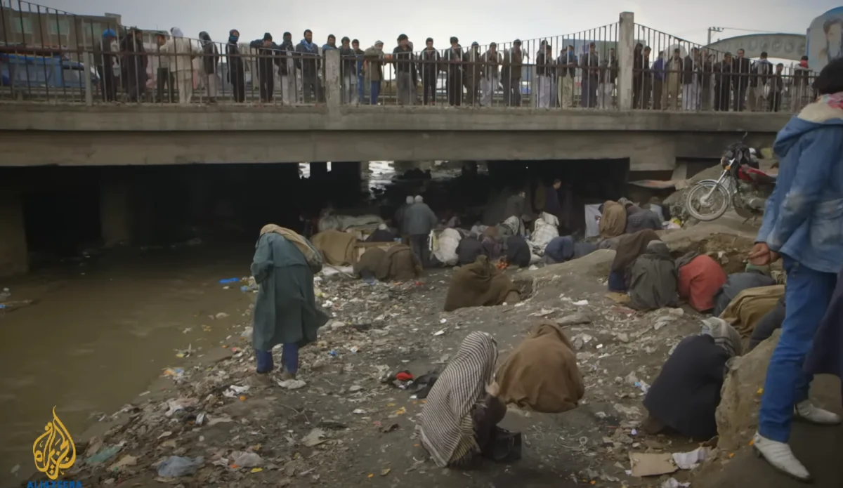 People gather on a bridge and along a debris-strewn riverbank beneath it, where others are huddled among the trash.