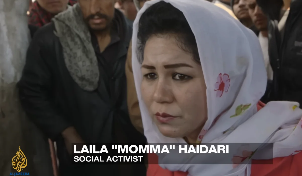 Laila "Momma" Haidari, an Afghan social activist, is shown in a close-up shot, wearing a white headscarf with floral patterns, amidst a crowd of people.