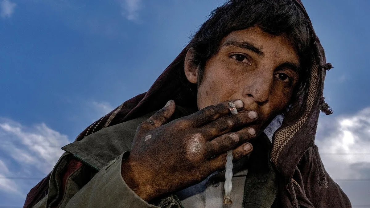A rugged-looking person with dirty hands smokes a cigarette, gazing intently. They wear a hooded jacket against a blue sky backdrop with clouds. They also appear to be holding drug paraphernalia, a plastic straw looking tube burnt at one end, implying they are smoking heroin.