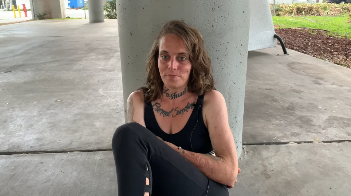 A woman with tattoos sits on the ground, leaning against a concrete pillar, for an interview to tell her addiction story. She has long brown hair, wearing a black tank top and leggings. The setting appears to be beneath an overpass, with a mix of concrete and greenery in the background. Her expression is neutral, with a slight closed-mouth smile.