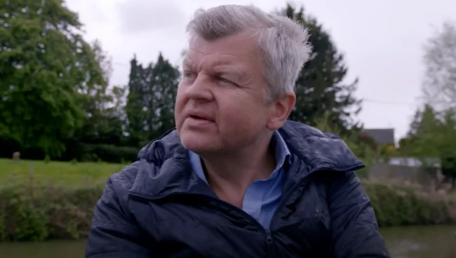 Adrian Chiles wearing a jacket sits on a boat, looking out over the water with a calm expression.
