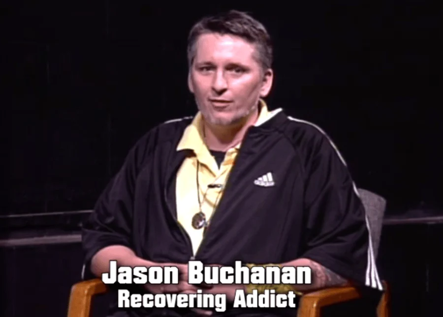 Jason Buchanan sits in a chair on a dark stage wearing a black Adidas jacket, with on-screen text identifying him as a recovering addict.