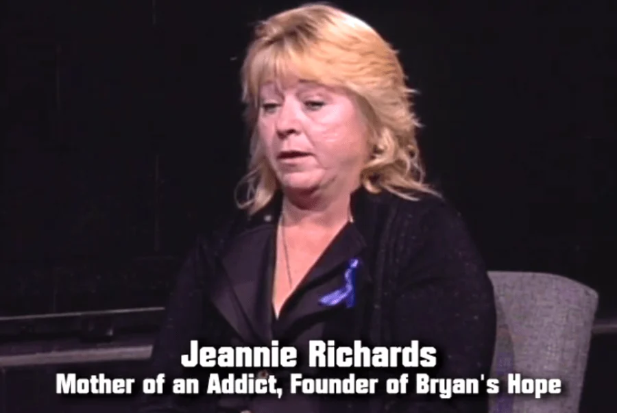 Jeannie Richards sits in a chair on a dark stage, with text identifying her as the mother of an addict and founder of Bryan’s Hope.