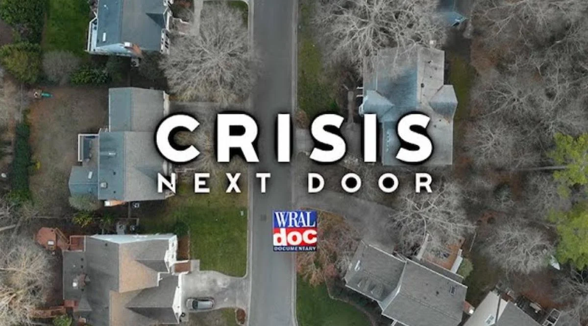 Aerial view of suburban homes with bare trees, centered text "Crisis Next Door" in bold, with a small "WRAL doc" logo below.