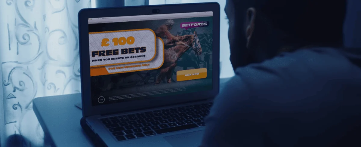 A man is sitting at a table with a laptop, browsing a betting website that offers 100 pounds in free bets.