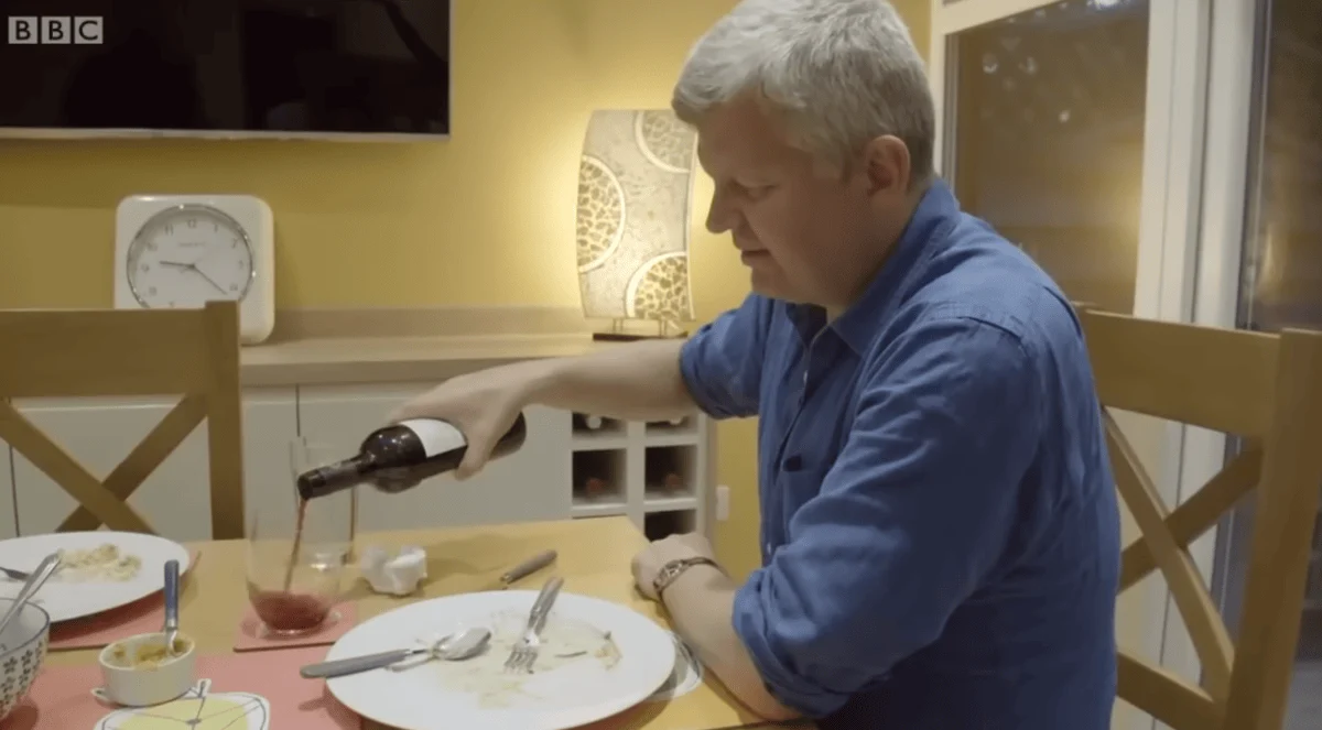 Adrian Chiles pouring red wine into a glass at a dining table