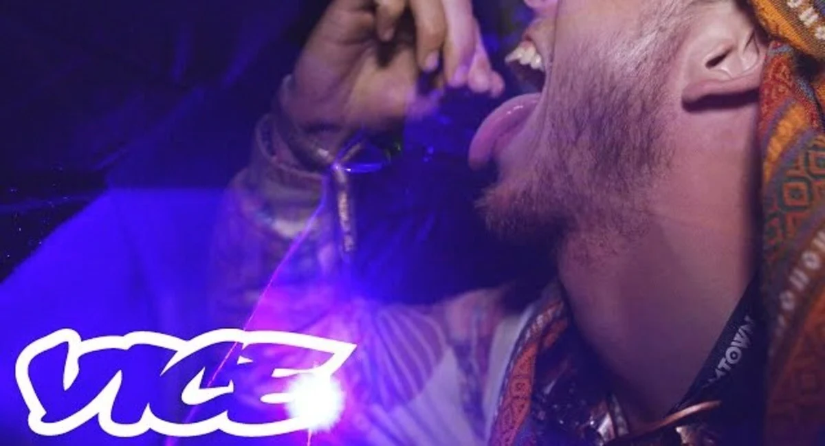 Close-up side view of a man with his mouth open and tongue out, placing an ecstasy pill in his mouth.