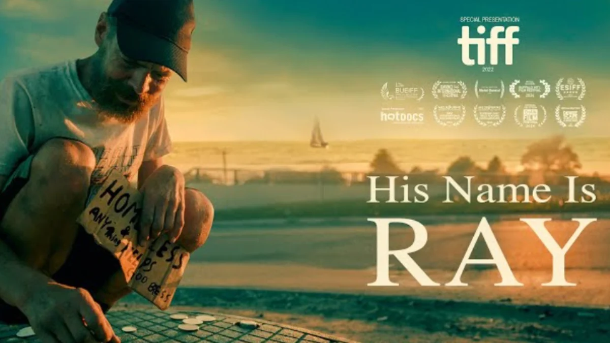 Ray sits on the ground with a "homeless" sign while counting change, with a background of the Toronto lakeshore, and His Name is Ray in big bold letters, alongside mention of multiple TIFF awards that the documentary received.
