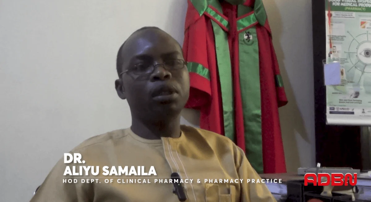 Dr. Aliyu Samaila, in a light brown shirt, sits in an office with a red and green academic gown hanging on the wall, exuding a professional tone.