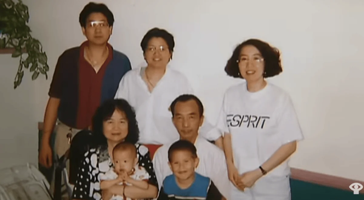 Group photo of a multi-generational East Asian family standing and sitting indoors in front of a white wall.