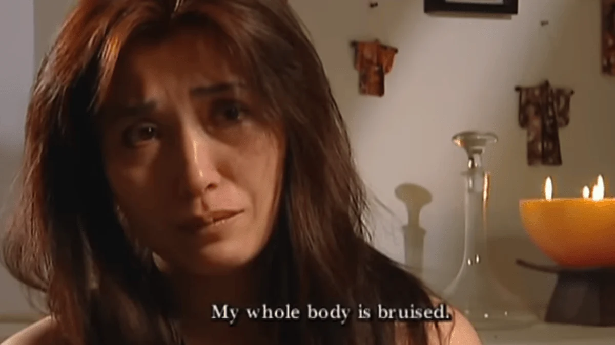 A close-up of a distressed, crying East Asian woman (Minh, Phillip’s ex-wife) with subtitle text reading, "My whole body is bruised."