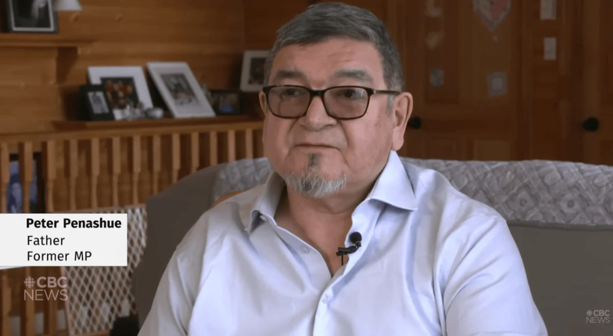 A man with glasses, wearing a light blue shirt, sits in a cozy room with wooden walls and family photos behind him. Text reads "Peter Penashue, Father, Former MP."