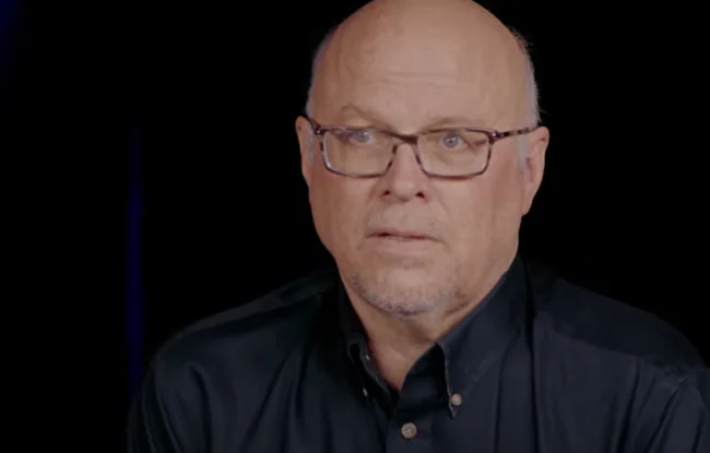 Close-up of Dr. Jeff Harries, a bald man in glasses, wearing a black shirt against a black background.