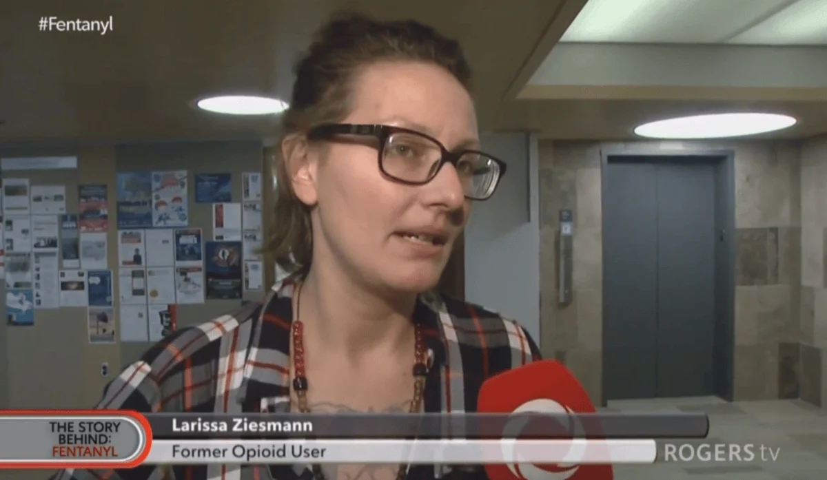 Woman wearing glasses speaks into a red microphone in a hallway. Chyron reads "Larissa Ziesmann, Former Opioid User." Neutral expression.
