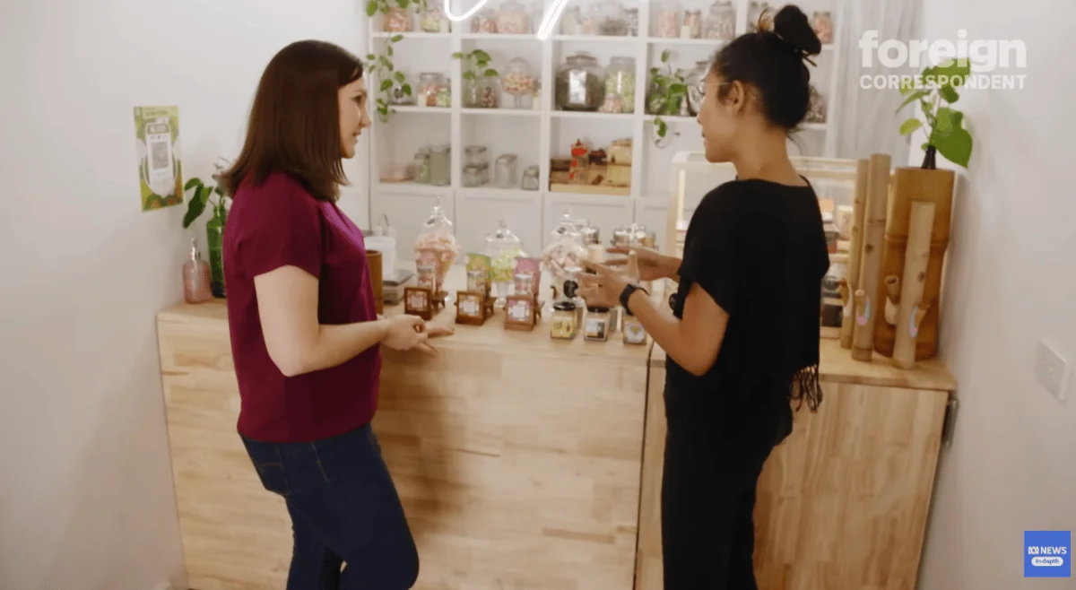 Two women stand in a cannabis shop, conversing. Shelves display jars of assorted cannabis-infused sweets behind them. The atmosphere is warm and inviting.