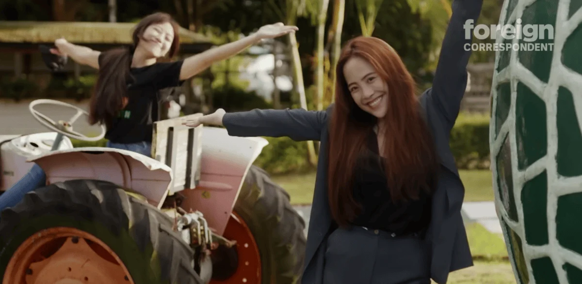 Two women joyfully posing outdoors; one on a pink tractor and the other beside a large sculpture. The mood is playful and lively.