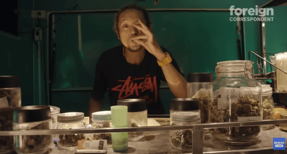 Man behind a counter in a dimly lit room, surrounded by jars of cannabis. He is holding a joint near his mouth and wearing a casual T-shirt and watch.