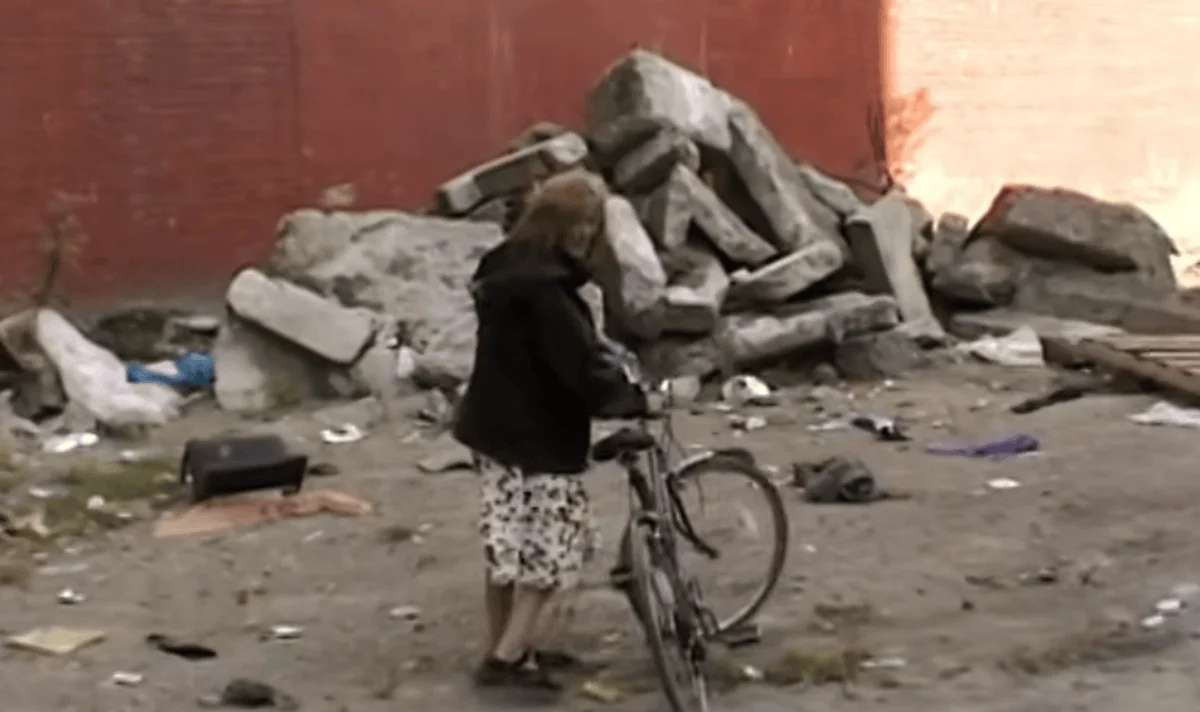 An older woman stands beside a bicycle on a littered street with a pile of large rocks against a red wall, conveying an urban, desolate atmosphere.