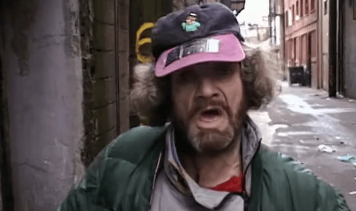 An older man with curly hair and a beard wears a cap and green jacket, standing in an empty alleyway. The scene conveys a gritty, urban atmosphere.