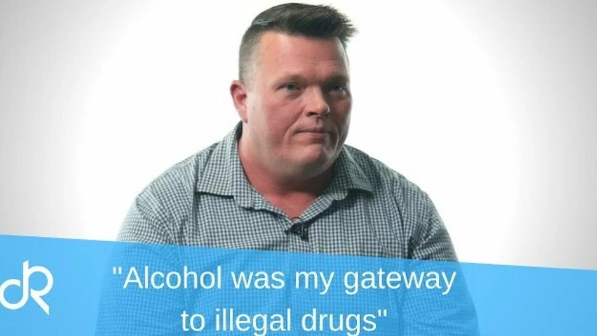 A man in a checkered shirt sits against a plain white background, looking contemplatively during his interview to share his addiction story. A blue banner reads, "Alcohol was my gateway to illegal drugs."
