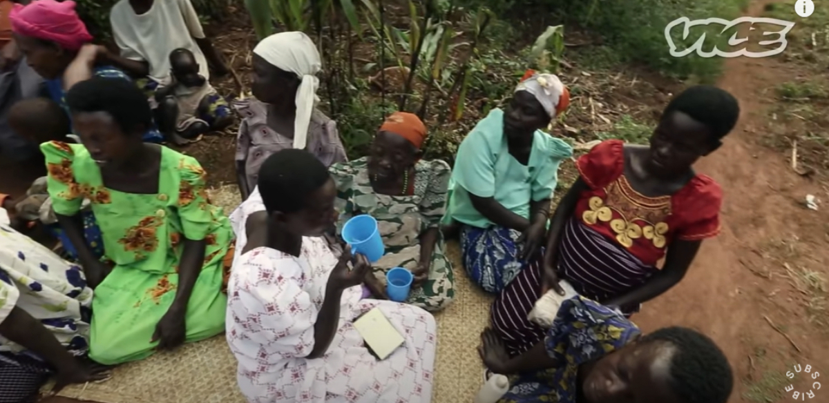 A group of Ugandan people seated on the ground in front of a tree, sharing drinks of waragi, a traditional moonshine beverage.