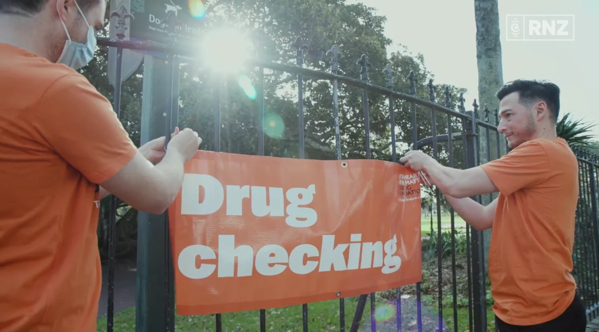 Two harm reduction workers in orange shirts display a "Drug Checking" sign on a fence to support community health initiatives.
