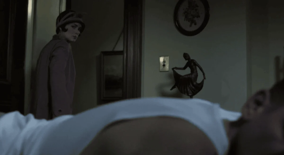 Lois Wilson in a cloche hat and coat stands in at their bedroom doorway, looking back at Bill sleeping in bed in the foreground.