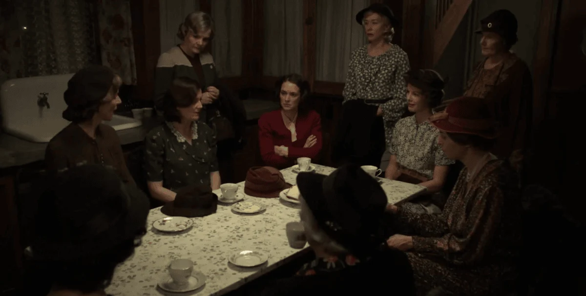 A group of women sitting and standing around a table with a floral tablecloth inside a dimly lit rustic room.